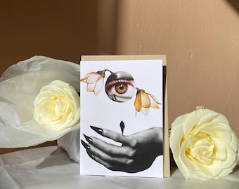 May include: Greeting card featuring a surrealist collage of an eye, flowers, and a hand with long black nails. The card is set against a backdrop of a white rose and a beige background, creating a striking contrast.