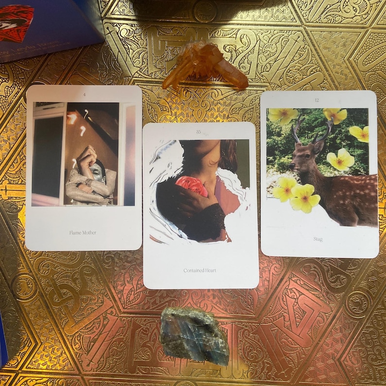 May include: Three tarot cards with images of a person, a deer, and a person holding a rose. The cards are on a gold patterned surface, with a crystal and a stone. The cards have the text "Flame Mother", "Contained Heart", and "Soul".