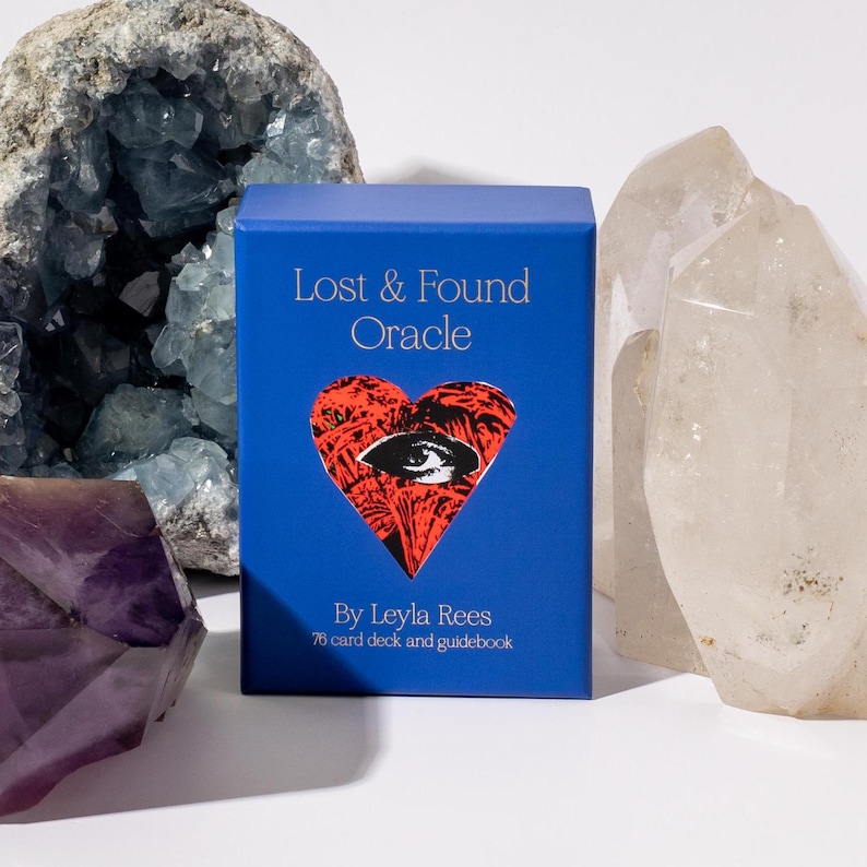 May include: A blue box for the "Lost & Found Oracle" by Leyla Rees, with a red heart design featuring an eye. The box is surrounded by crystals, including amethyst and quartz. The text on the box reads "78 card deck and guidebook."