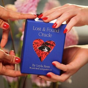 May include: A blue box for the "Lost & Found Oracle" deck, held by hands with red nail polish. The box features a red heart with an eye design. Text on the box reads "By Leyla Rees" and "78 card deck and guidebook."