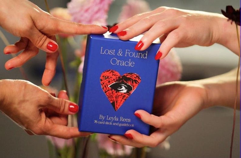 May include: A blue box for the "Lost & Found Oracle" deck, held by hands with red nail polish. The box features a red heart with an eye design. Text on the box reads "By Leyla Rees" and "78 card deck and guidebook."