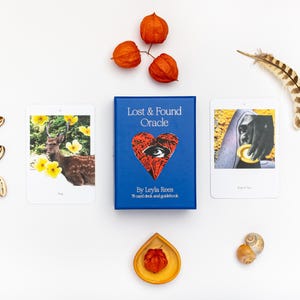 May include: A blue box set of the "Lost & Found Oracle" by Leyla Rees, with two oracle cards and decorative elements. The cards feature images of a deer and a person. The set includes a guide book and is surrounded by orange flowers, shells, and a feather.