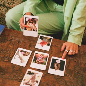 May include: A collection of tarot cards with various images, including hands, flowers, and a leopard, are arranged on a dark wooden table. A person in a light green suit holds a card, while another hand points to a card. The cards appear to be part of a divination set.