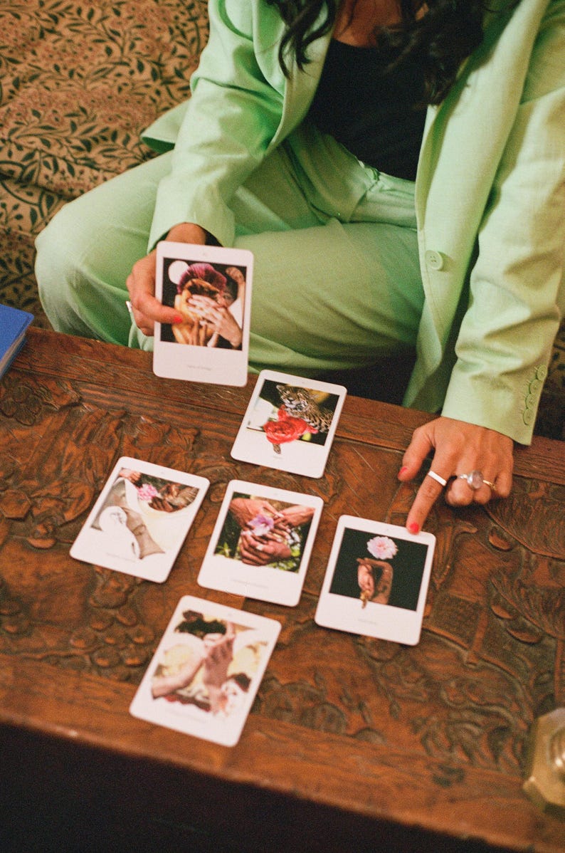 May include: A collection of tarot cards with various images, including hands, flowers, and a leopard, are arranged on a dark wooden table. A person in a light green suit holds a card, while another hand points to a card. The cards appear to be part of a divination set.