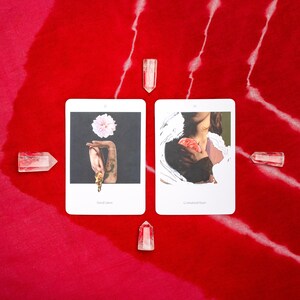 May include: Two tarot cards with illustrations and text, surrounded by clear quartz crystals, are displayed on a red fabric with white streaks. One card shows a hand holding a flower, the other a person holding a heart.