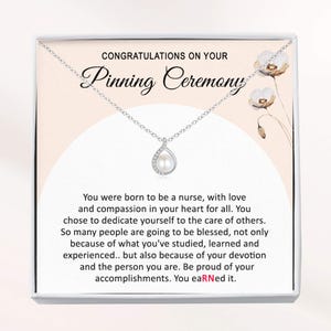 May include: A silver necklace with a pear-shaped pendant containing a pearl and surrounding crystals. The necklace is displayed in a white box with text that reads "CONGRATULATIONS ON YOUR Pinning Ceremony."