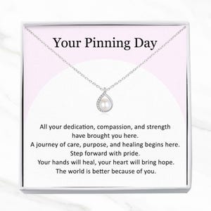May include: A silver necklace with a teardrop pendant featuring a pearl and small clear stones. The necklace is presented in a white box with the text "Your Pinning Day" and inspirational text.