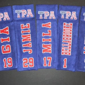 May include: Seven blue towels with white and red text. Each towel has a name and number. The top of each towel has the letters "TPA" in a patriotic font. Names include Kinsey, Gia, Jamie, Mila, Bellabrooke, Aleena, and Eva.