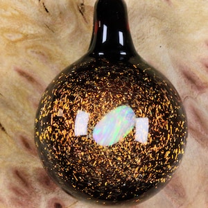 May include: A handcrafted glass pendant with a black base and a speckled gold design. The pendant features a central oval of iridescent white and green, and a black top. The pendant is approximately 2 inches in length.