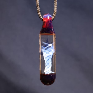 May include: A glass pendant necklace with a swirling blue and white design encased in a clear, rectangular glass bottle shape. The top is red and purple, and it hangs from a gold chain.