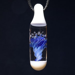 Puede incluir: Un colgante de cristal transparente con un diseño de remolino azul suspendido en líquido. El colgante tiene una parte superior e inferior blancas y está suspendido de una cadena plateada. El fondo es azul oscuro.