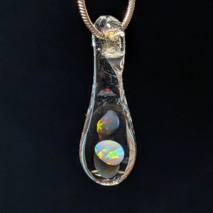 Peut inclure: Un pendentif en verre transparent avec trois opales à l'intérieur, suspendu à une chaîne argentée. Les opales affichent des couleurs irisées, dont le jaune, le vert et le bleu, sur fond sombre. Le pendentif est en forme de goutte.