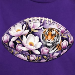 May include: A purple t-shirt featuring a football design with a tiger's face surrounded by white and purple flowers. The design is centered on the shirt, creating a bold and eye-catching look. The shirt is made of a soft material.