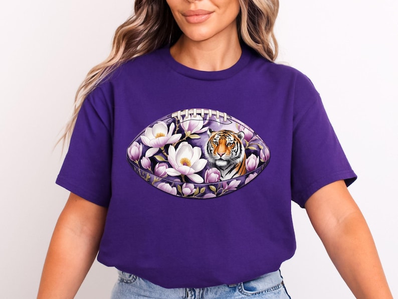 May include: A purple t-shirt featuring a football design with a tiger's face surrounded by white and purple flowers. The design is centered on the shirt, creating a bold and eye-catching look. The shirt is made of a soft material.