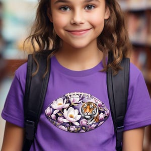 Pode incluir: Uma camiseta roxa com um design de futebol americano com flores brancas, detalhes roxos e o rosto de um tigre. A menina usa uma mochila preta. O fundo &eacute; uma estante desfocada.