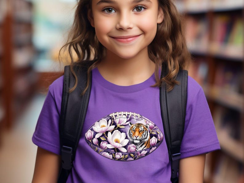 Pode incluir: Uma camiseta roxa com um design de futebol americano com flores brancas, detalhes roxos e o rosto de um tigre. A menina usa uma mochila preta. O fundo &eacute; uma estante desfocada.