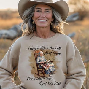 May include: Beige sweatshirt with the text "I Used to Talk to my Kids About Jesus Now I Talk to Jesus 'Bout My Kids". The graphic depicts an elderly woman in a rocking chair reading a book. The woman in the image is wearing a cowboy hat.