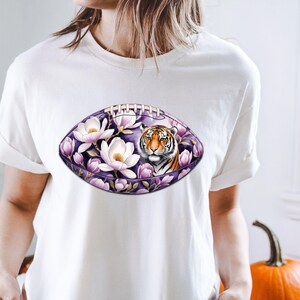 Pode incluir: Camiseta branca com um design em forma de bola de futebol americano. O design apresenta o rosto de um tigre rodeado por flores brancas e p&eacute;talas roxas. O fundo &eacute; roxo escuro.