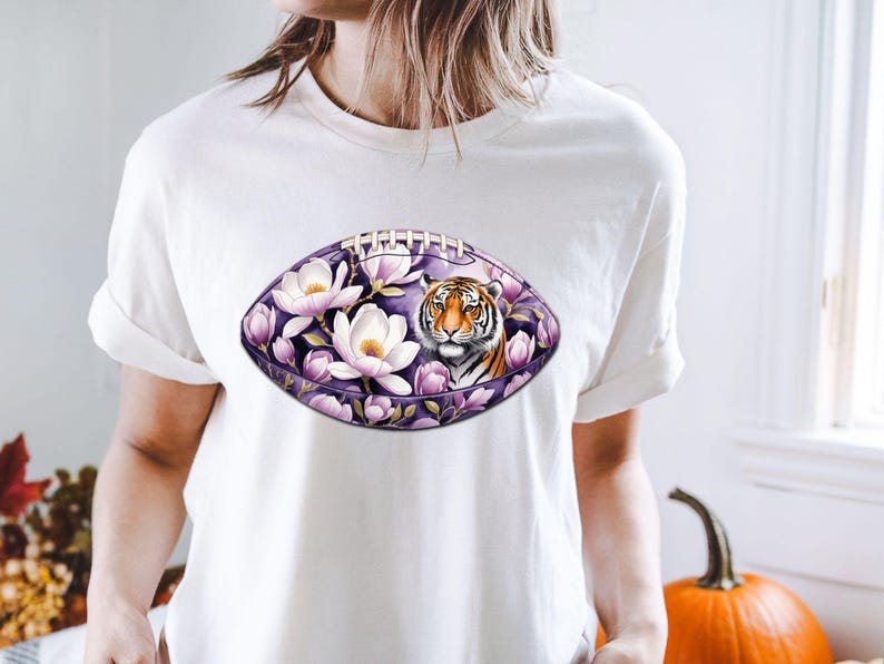 Pode incluir: Camiseta branca com um design em forma de bola de futebol americano. O design apresenta o rosto de um tigre rodeado por flores brancas e p&eacute;talas roxas. O fundo &eacute; roxo escuro.