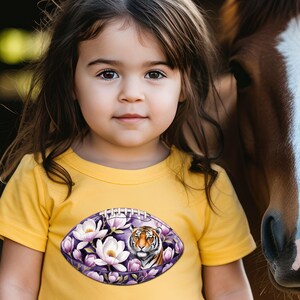 Pode incluir: Uma crian&ccedil;a usa uma camiseta amarela com um desenho em forma de bola de futebol. O desenho apresenta um tigre rodeado por flores brancas e roxas. Um cavalo marrom est&aacute; ao lado da crian&ccedil;a.