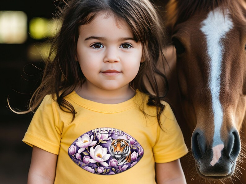 Pode incluir: Uma crian&ccedil;a usa uma camiseta amarela com um desenho em forma de bola de futebol. O desenho apresenta um tigre rodeado por flores brancas e roxas. Um cavalo marrom est&aacute; ao lado da crian&ccedil;a.