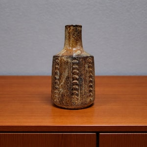 May include: A brown and tan ceramic vase with a textured, vertical pattern. The vase has a narrow neck and a wider body, standing on a wooden surface. The vase is approximately 8 inches tall.
