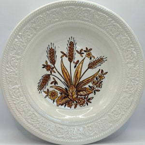 May include: White ceramic dinner plate with a raised embossed border design. The center of the plate features a brown and yellow floral design with wheat stalks.