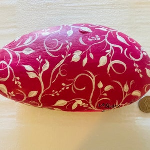 Puede incluir: Una piedra pintada de rosa con un dise&ntilde;o floral blanco. La piedra tiene forma ovalada y tiene la firma "LA Stanley" en la parte inferior.