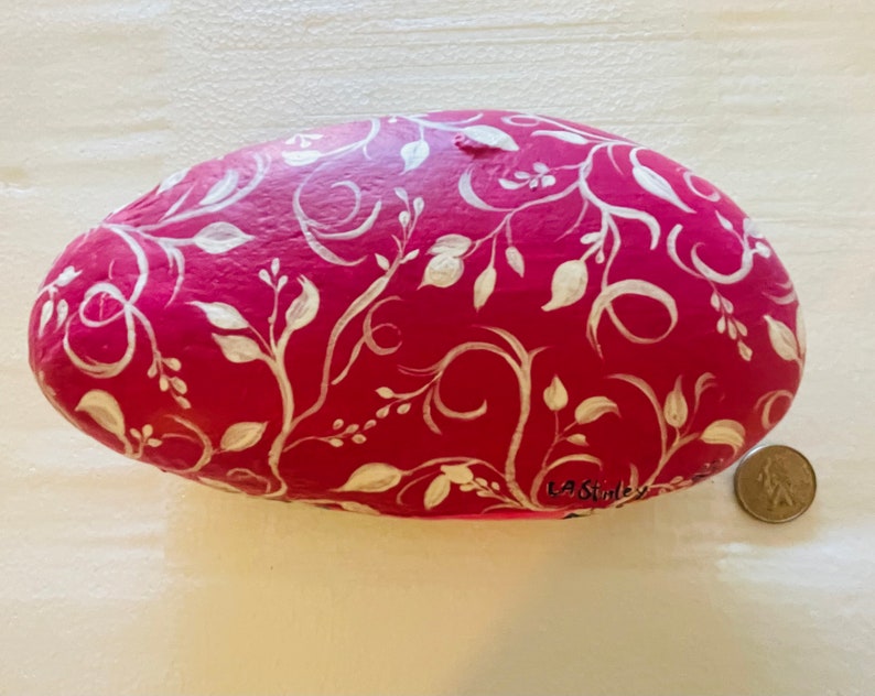 Puede incluir: Una piedra pintada de rosa con un dise&ntilde;o floral blanco. La piedra tiene forma ovalada y tiene la firma "LA Stanley" en la parte inferior.