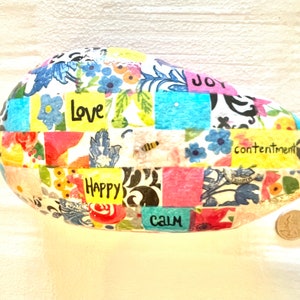 Puede incluir: Una roca ovalada blanca con un dise&ntilde;o de patchwork de patrones florales coloridos y cuadrados. La roca tiene las palabras "Love", "Happy", "Calm", "Joy" y "Contentment" escritas en ella.