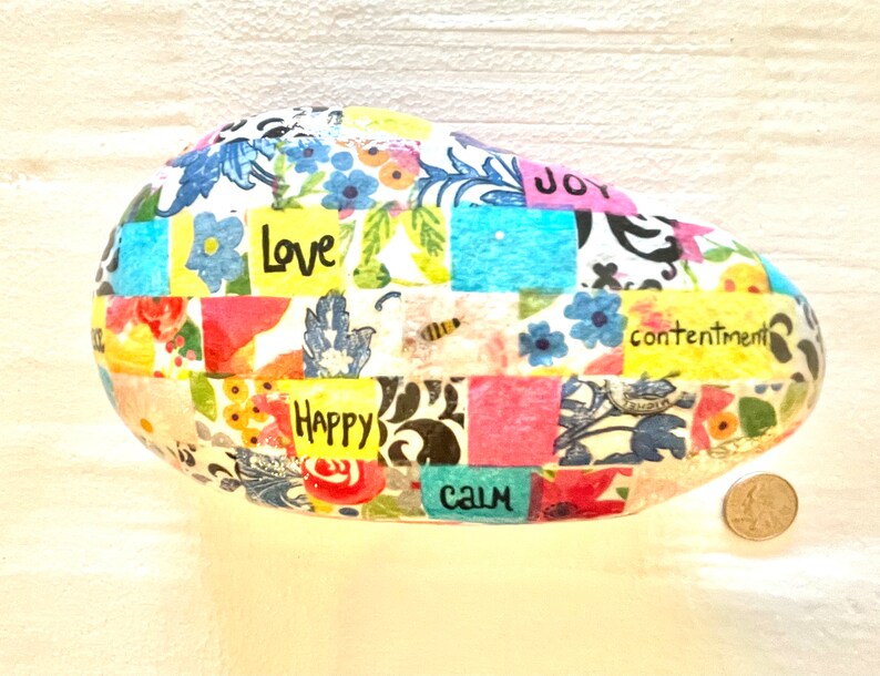 Puede incluir: Una roca ovalada blanca con un dise&ntilde;o de patchwork de patrones florales coloridos y cuadrados. La roca tiene las palabras "Love", "Happy", "Calm", "Joy" y "Contentment" escritas en ella.