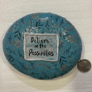 May include: A round, blue paperweight with a white floral pattern. The paperweight has a white square with the text "Believe in the Possibilities" written in black.