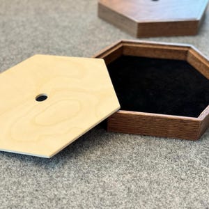 May include: A wooden hexagonal box with a removable lid. The lid is made of light wood with a natural grain pattern. The box is lined with black felt and has a small hole in the center of the lid.