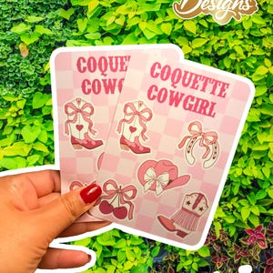 Puede incluir: Dos pegatinas rosas y blancas con temática vaquera. Las pegatinas presentan ilustraciones de una bota de vaquero, una herradura, un sombrero y cerezas. El texto "COQUETTE COWGIRL" está impreso en las pegatinas.