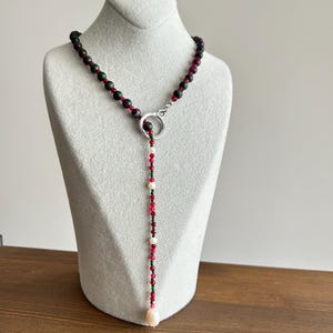 Peut inclure: Un collier de perles en forme de Y. Il présente des perles rouge foncé, vertes et blanches, avec un fermoir en forme d'anneau argenté. Le pendentif se termine par une grosse perle en forme de goutte d'eau rose clair. Le collier est exposé sur un mannequin blanc.