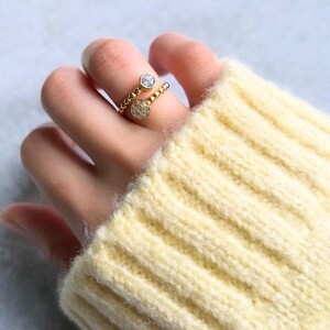 Peut inclure: Une bague dorée ornée de deux pierres précieuses rondes et transparentes. La bague présente un motif de perles sur le corps. La bague est portée au doigt, et la personne porte un pull jaune.