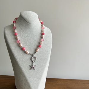 May include: A necklace with pink and white beads, and a silver starfish pendant. The necklace is displayed on a gray mannequin. The beads are a mix of round and oval shapes, with silver spacers.