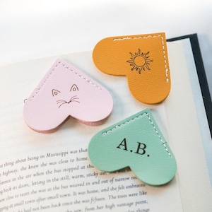 May include: Three heart-shaped leather bookmarks with different designs. The first bookmark is pink with a cat face, the second is orange with a sun, and the third is green with the initials "A.B."
