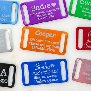 May include: A collection of colorful pet ID tags in various colors, including blue, purple, green, red, silver, black, and pink. Each tag features a pet's name and contact information, with some including additional text.
