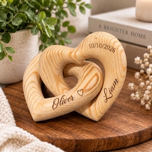 Puede incluir: Dos corazones de madera entrelazados tallados en madera de color claro con vetas visibles. Los corazones están grabados con los nombres Oliver y Liam, un pequeño símbolo de corazón y la fecha 10/10/2026 en letra cursiva y de imprenta marrón.