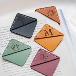 May include: A collection of leather bookmarks in various colors, including black, orange, green, brown, and pink. Each bookmark features a unique embossed design: a cross, the letter "M", a paw print, a sun, and a flower. White stitching outlines each bookmark.