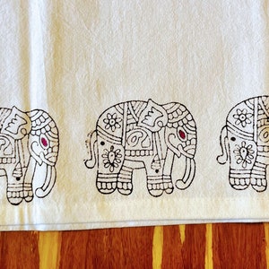 May include: White cotton fabric with a black and pink patterned border of five elephants. The elephants are intricately designed with floral motifs and have pink accents on their trunks.