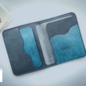 Op de afbeelding: Een leren portemonnee met twee vouwen in grijstinten en blauwgroen. De portemonnee heeft meerdere kaartsleuven en een biljettenvak. Het woord "SARETS" is zichtbaar op de voorkant. Een kleine kaart met een handafdruk staat op de achtergrond.