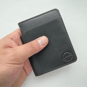 Könnte beinhalten: Ein schwarzer Kartenhalter aus Leder mit minimalistischem Design. Das Portemonnaie hat ein schlankes Profil mit mehreren Kartenfächern und einem abgesteppten Rand. Ein kreisförmiges Logo ist in der unteren rechten Ecke geprägt. Der Kartenhalter wird in einer Hand gehalten.