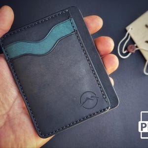 Puede incluir: Un tarjetero de cuero negro con un acento verde azulado y bordes cosidos. El tarjetero presenta un logotipo de montaña y se sostiene en una mano. Un icono PDF está en la esquina inferior derecha.