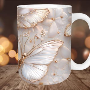 May include: White ceramic mug featuring a 3D butterfly and floral design. The butterfly has white wings with gold accents and is adorned with small crystals. The background has a soft, blurred effect.