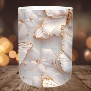 May include: A white ceramic mug featuring a repeating pattern of white flowers and butterflies with gold accents and embedded crystals. The design is embossed, creating a 3D effect. The mug has a smooth, glossy finish.