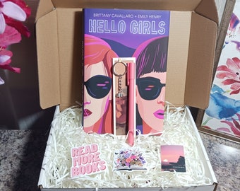 May include: A white box with a brown lid is open and filled with white packing peanuts. Inside the box is a pink and purple book titled "Hello Girls" by Brittany Cavallaro and Emily Henry. There is also a pink pen, a keychain with a key and a charm, a sticker that says "Read More Books", and a sticker with a sunset scene.