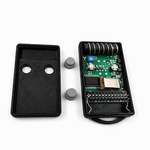 May include: An open black electronic device with a green circuit board, various components, and a black cover with two holes. Two gray buttons and a detached black cover are also present.