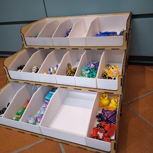 Può includere: Un organizer in legno a tre livelli con scomparti bianchi, riempiti con vari peluche. L'organizer ha una cornice marrone chiaro ed è progettato per contenere piccoli oggetti, mostrando una collezione di animali di peluche colorati.